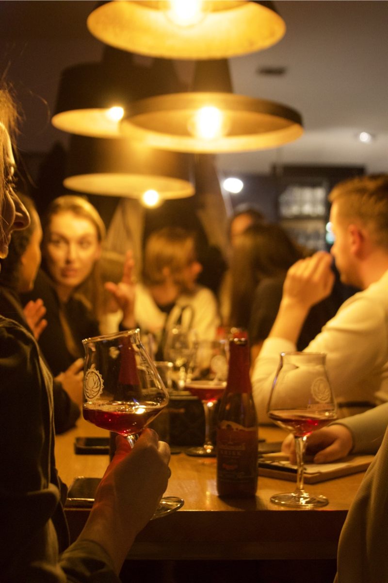 Gäste sitzen abends unter warmen Lampen am langen Tisch, im Vordergrund Gläser mit dunklem Bier