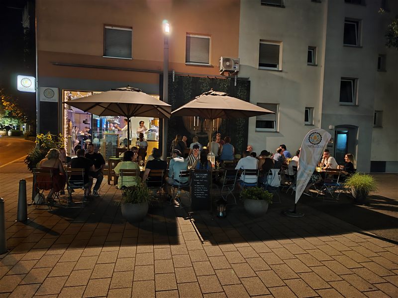 Volle Terrasse vor dem Craftelicious an einem Sommerabend, Gäste sitzen unter Sonnenschirmen vor dem Hopfen-Wandbild